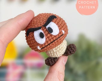 May include: A crocheted Goomba character from the Super Mario Bros. video game series. The brown mushroom-shaped figure has a stern expression with black eyebrows, white eyes, and a small mouth. The text "CROCHET PATTERN" is in a pink circle.