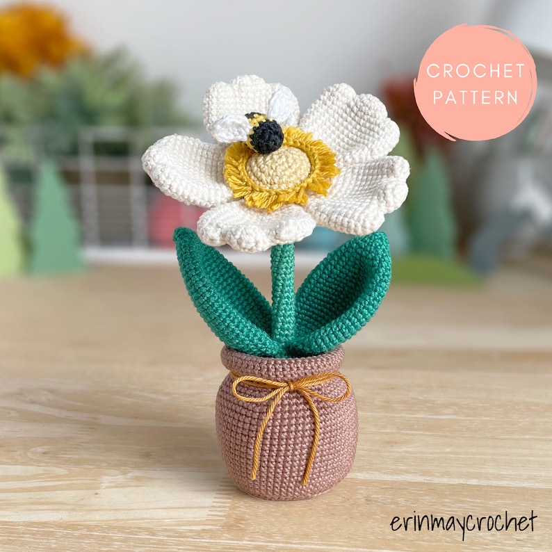 May include: Crochet pattern for a white flower with a yellow center and a bee, in a brown pot with a green stem and leaves. The pattern is called "Crochet Pattern".