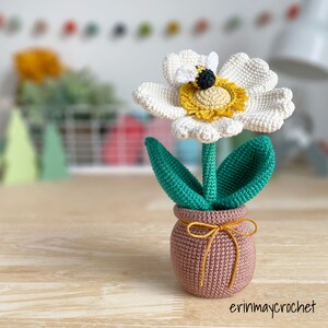 May include: A crocheted white flower with a yellow center and a bee on top, in a pink crocheted pot with a brown ribbon.
