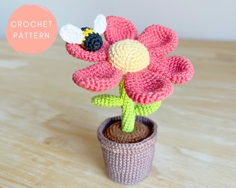 May include: A crocheted flower with a pink center and yellow petals, a green stem, and a brown pot. A crocheted bee is perched on the flower.