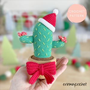 May include: Crochet pattern for a green cactus wearing a red and white Santa hat with a red bow on the pot. The cactus has two pink flowers.