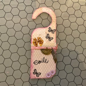 Könnte beinhalten: Ein rosa Türklopfer mit weißer Spitzenborte und einer braunen Schleife. Der Türklopfer hat oben einen Haken und zeigt ein Schmetterlingsdesign, eine fächerförmige Verzierung und das Wort "Smile" in Schwarz.