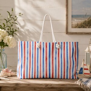 May include: A rectangular tote bag featuring a vertical striped pattern in watercolour-style red and blue on a white background. The bag has two thick white rope handles attached with knots through metal grommets at the top of the bag.