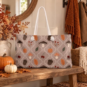 May include: A tote bag with a light brown background featuring a repeating pattern of orange pumpkins, brown acorns, red berries, green leaves, and various mushrooms. The bag has two white rope handles attached with silver metal grommets.