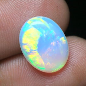 May include: An oval-shaped cabochon opal gemstone featuring a translucent white base with vibrant flashes of yellow, green, and blue light play across its polished, smooth, and domed surface, showcasing a distinct iridescent colour display.