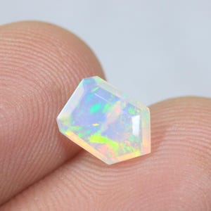 Peut inclure: Une pierre précieuse opale à facettes en forme de bouclier, présentant une base blanc laiteux translucide avec des éclats vibrants de vert néon, de jaune et d'orange sur toute sa surface, avec une finition polie et une coupe géométrique.