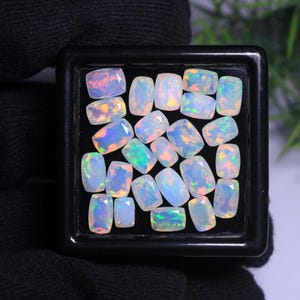 May include: A collection of twenty-one small, rectangular, faceted opals arranged in a black display box. Each gemstone exhibits a translucent, milky white base with vibrant flashes of iridescent green, orange, and blue color play throughout.