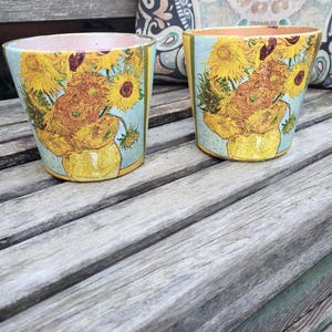 May include: Two ceramic flower pots with a light blue background and a yellow border. The pots feature a print of Van Gogh's "Sunflowers" painting. The pots are sitting on a weathered wooden surface.