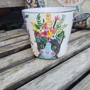 May include: A white ceramic flower pot with a cat face design and a colorful floral arrangement. The pot has a pink interior and a silver rim. The cat has green eyes and a blue bow.