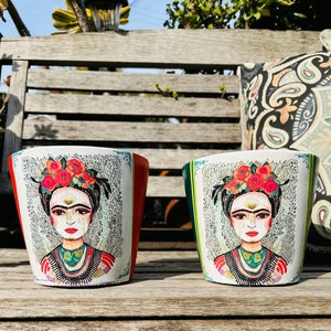 May include: Two white ceramic flower pots with colorful illustrations of Frida Kahlo. The pots have a green and red trim.