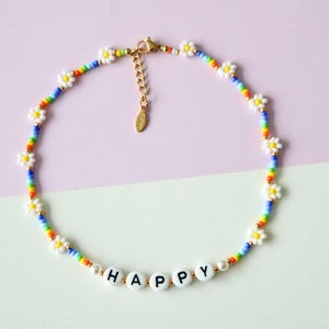 Könnte beinhalten: Eine Perlenkette mit einem regenbogenfarbenen Strang und weißen Gänseblümchen-Akzenten. Die Halskette trägt das Wort "HAPPY" in weißen Buchstabenperlen. Der Verschluss ist goldfarben.