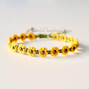 May include: A handmade bracelet with a repeating pattern of yellow crocheted flowers with brown centres. The bracelet is strung on a green cord and is displayed on a white surface. The text "Land L handmade store Etsy" is visible.