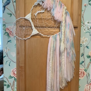 May include: A whimsical unicorn dreamcatcher with a white horn, ears, and face. The dreamcatcher features a woven web design and is adorned with pastel-colored yarn and fabric. It hangs on a wooden cabinet against a floral wallpaper background.