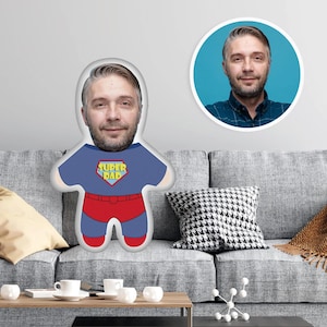 May include: A blue and red superhero cushion with the words "Super Dad" on the chest. The cushion has a person's face on it.
