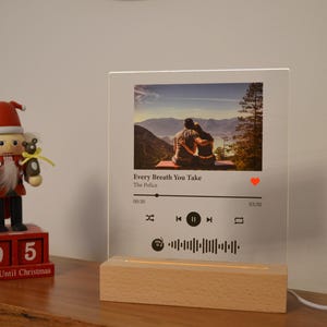 May include: A clear acrylic photo frame with a wooden base. The frame displays a photo of a couple in a woodland setting. The frame also includes a Spotify music player with the song "Every Breath You Take" by The Police playing.