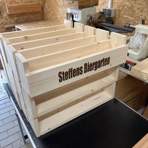 May include: A wooden shelf unit with three shelves, made of unfinished pine wood. The shelf unit has the words "Steffens Biergarten" engraved on the front.