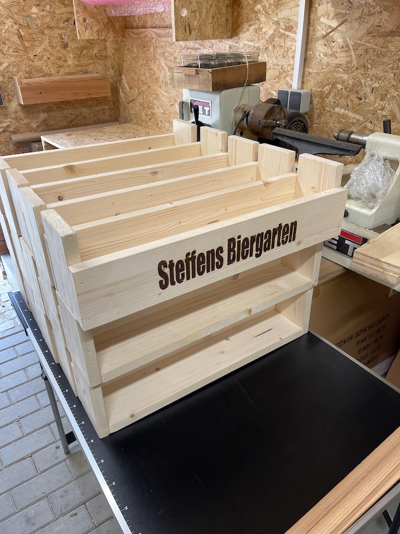May include: A wooden shelf unit with three shelves, made of unfinished pine wood. The shelf unit has the words "Steffens Biergarten" engraved on the front.