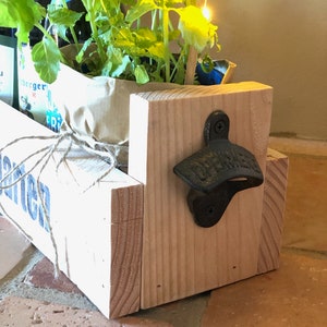 May include: A wooden planter box with a white painted finish and the word "Garten" printed on the front. The planter box has a black metal bottle opener attached to the side. The planter box is filled with green plants.