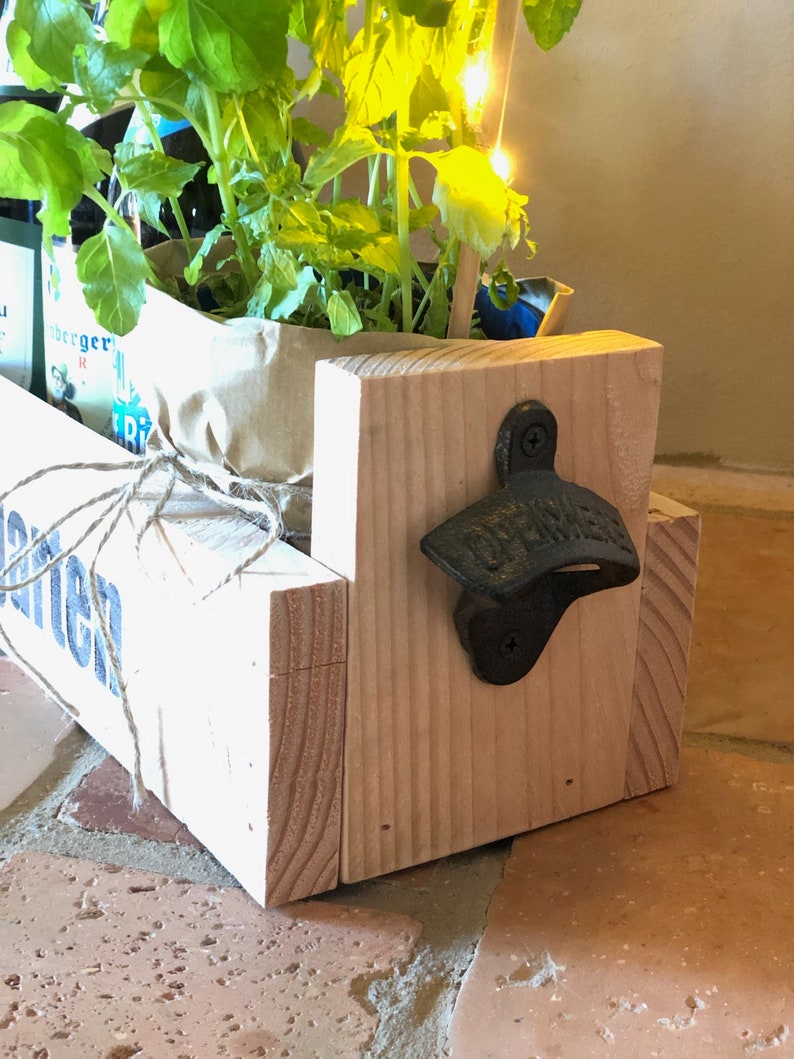 May include: A wooden planter box with a white painted finish and the word "Garten" printed on the front. The planter box has a black metal bottle opener attached to the side. The planter box is filled with green plants.
