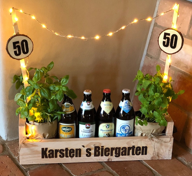 May include: A wooden crate with the words "Karsten's Biergarten" on the front. The crate is decorated with string lights and two wooden signs with the number 50 on them. There are four bottles of beer in the crate, and two potted plants.