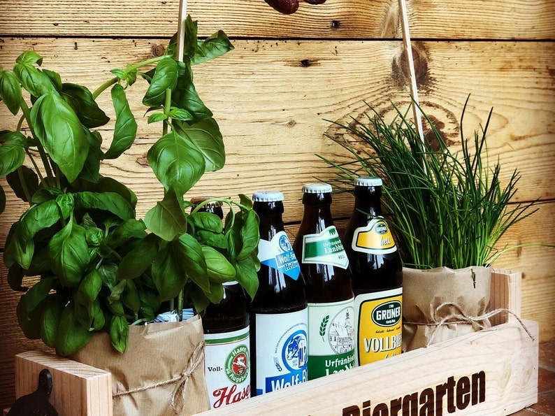 May include: A wooden crate with the word "Biergarten" engraved on the front. The crate contains four bottles of beer, two potted basil plants, and one potted chives plant.