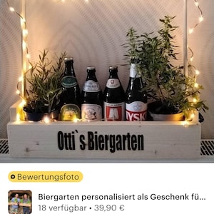 May include: A wooden crate with a sign that reads "Otti's Biergarten" and is decorated with string lights and potted plants. There are six bottles of beer in the crate.