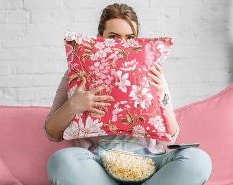May include: A red floral patterned pillow with white and pink flowers is held up in front of a person's face. The person is sitting on a pink couch with a bowl of popcorn and a remote control.