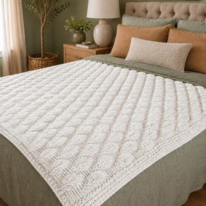Può includere: Una coperta bianca a maglia a trecce, drappeggiata su un letto. La coperta presenta un motivo a rombi ripetuto e un bordo strutturato. Il letto è fatto con lenzuola verdi e diversi cuscini nei toni del marrone e del beige.