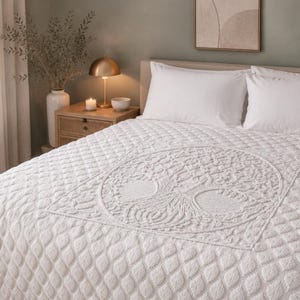 May include: A white bedspread with a tree of life design in the center. The bedspread has a textured, quilted pattern. Two white pillows rest on the bed. A nightstand with a lamp and candles is visible.
