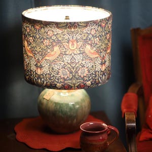May include: A decorative table lamp with a patterned lampshade featuring birds and floral designs. The lamp sits on a green ceramic base. A red mug and a red placemat are in the foreground, with a red chair in the background.