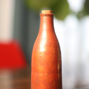 Pode incluir: Uma garrafa alta, castanho-alaranjada, com uma rolha de cortiça. A garrafa tem um gargalo fino e um corpo arredondado. O fundo está desfocado, com toques de vermelho e verde. Esta garrafa decorativa é adequada para decoração de casa.