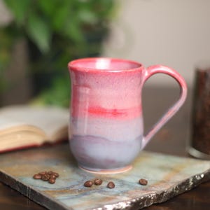 May include: A ceramic mug featuring a smooth gradient glaze that transitions from a vibrant pink at the rim to a soft lavender and light blue at the base. The mug has a curved body and a single handle, showcasing a glossy finish throughout.
