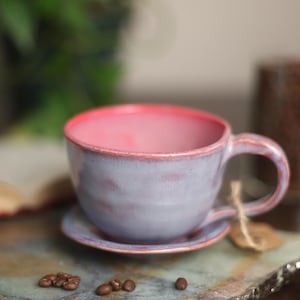 May include: A ceramic mug and matching saucer set featuring a light lavender exterior and a vibrant pink interior. The glaze has a mottled, textured appearance with pink rim accents. A small brown paper tag is tied to the handle with twine.