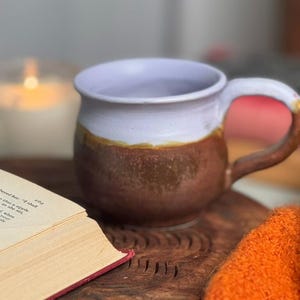 May include: A handmade ceramic mug with a lavender rim, a brown body, and a brown handle. The mug sits on a wooden surface next to an open book and an orange textile. A lit candle is in the background.