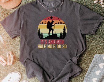 Op de afbeelding: Een antracietgrijs T-shirt met korte mouwen en een afbeelding van een wandelaar met een rugzak en wandelstok op een bergtop. Het ontwerp bevat een zonsondergang met horizontale strepen en de tekst IT'S ANOTHER HALF MILE OR SO.