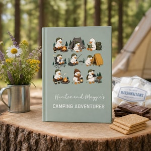 May include: A light sage green hardback book titled Hunter and Maggie's CAMPING ADVENTURES, featuring a grid of nine illustrations of cartoon ducks engaged in various camping activities like roasting marshmallows, fishing, and reading.