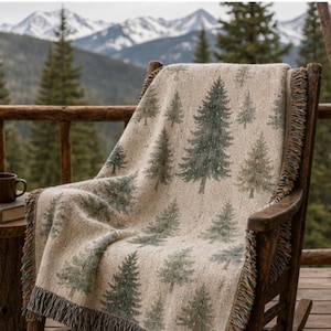 May include: A beige throw blanket with a repeating pattern of green pine trees draped over a wooden rocking chair. The blanket has a fringed edge and is set against a backdrop of mountains and evergreen trees.