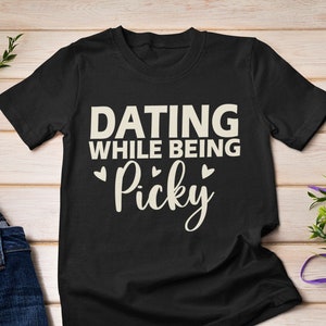 Può includere: Maglietta nera con il testo "Dating while being Picky" in lettere bianche con tre piccoli cuori.