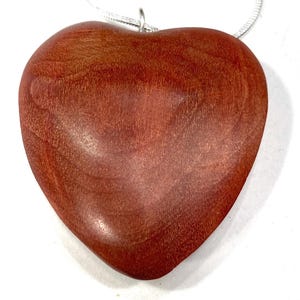 May include: A heart-shaped pendant made of reddish-brown wood. The pendant is smooth and polished, with visible wood grain. It hangs from a thin, silver-colored chain, creating a simple and elegant necklace.