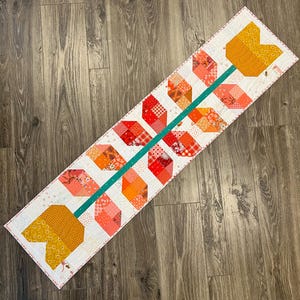 May include: A quilted table runner with a floral arrow design. The arrow features a teal stem and multi-colored flower petals in shades of orange, red, and yellow. The runner is bordered with a white fabric and a pink stitched edge.