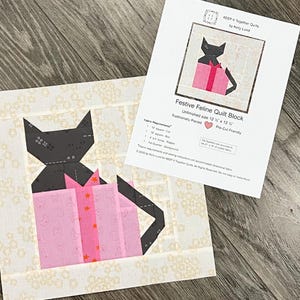 Könnte beinhalten: Ein Stoff-Quiltblock mit einer schwarzen Katze und einer rosa Geschenkbox. Der Block befindet sich auf einem cremefarbenen Hintergrund mit einem Blumenmuster. Ein Papier mit dem Text "Festive Feline Quilt Block" liegt neben dem Block. Unfertige Größe 31,75 cm x 31,75 cm.