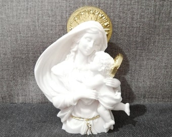 May include: White ceramic figurine of the Virgin Mary holding a baby Jesus. The figurine has a gold halo and is on a gray background.