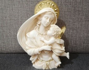 May include: A white ceramic sculpture of the Virgin Mary holding a baby Jesus. The sculpture has a gold halo and a gold trim around the Virgin Mary's robe.