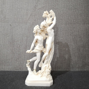 Könnte beinhalten: Weiße Marmorskulptur von zwei sich umarmenden Figuren, eine hält einen Zweig mit Blättern, auf einem weißen Sockel.