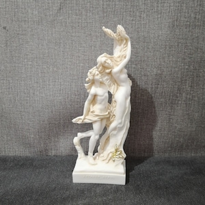 Könnte beinhalten: Weiße Marmorskulptur von zwei sich umarmenden Figuren, eine steht hoch und die andere streckt die Arme nach oben, auf einem weißen Sockel mit goldenen Akzenten.