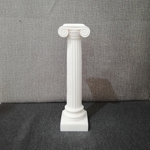 Puede incluir: Un pedestal de cerámica blanca con una columna estriada y una base cuadrada. La parte superior del pedestal tiene un diseño decorativo de volutas.