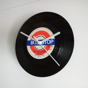 Puede incluir: Un reloj de vinilo negro con agujas blancas. La etiqueta del disco es roja, blanca y azul con el texto "BUS STOP" y "BILLY - DON'T BE A BEAR".