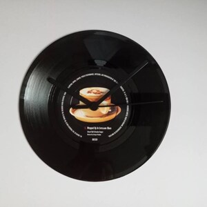 Puede incluir: Un reloj de vinilo negro con manecillas negras. La etiqueta del disco presenta una imagen de una taza de café y el texto "Wrapped Up in Lonesome Blues". El reloj está montado en una pared blanca.
