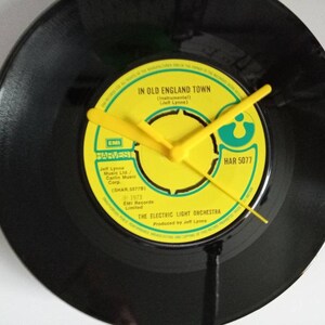 Puede incluir: Un reloj de disco de vinilo negro con manecillas amarillas. La esfera del reloj es una etiqueta amarilla con el texto "IN OLD ENGLAND TOWN" y "THE ELECTRIC LIGHT ORCHESRA". Las manecillas son amarillas.