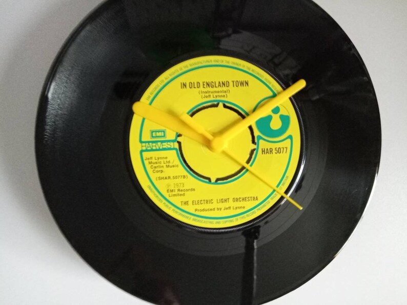 Puede incluir: Un reloj de disco de vinilo negro con manecillas amarillas. La esfera del reloj es una etiqueta amarilla con el texto "IN OLD ENGLAND TOWN" y "THE ELECTRIC LIGHT ORCHESRA". Las manecillas son amarillas.